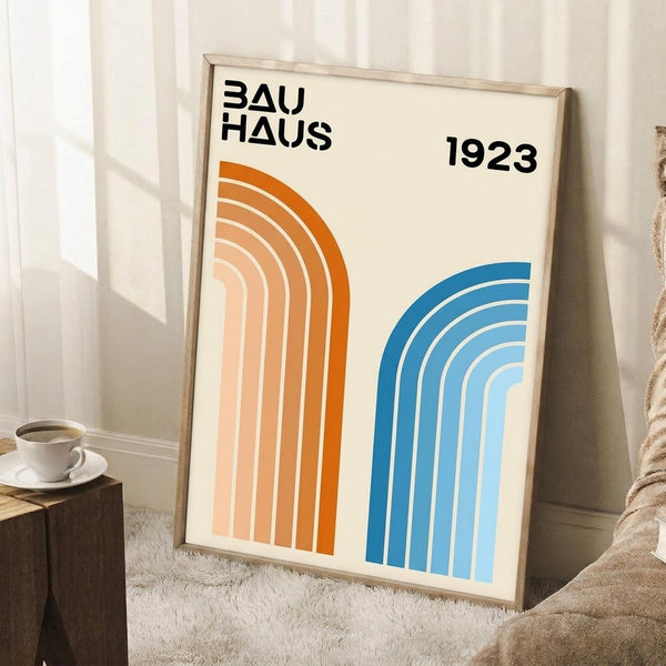 May include: Framed geometric poster with "BAUHAUS 1923" text and curved rainbow stripes in orange and blue gradients, displayed on white carpet against white paneled wall. Light wood frame, positioned next to wooden side table with white coffee cup and saucer. Beige knit throw blanket visible on right side. Natural lighting creates soft shadows.