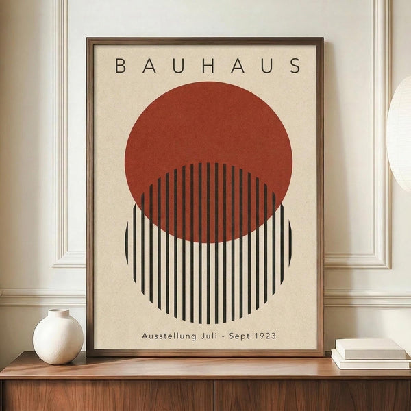 May include: Framed Bauhaus exhibition poster with red circle overlapping black and white striped circle on cream background, displayed on wooden sideboard next to white ceramic vase and books in room with white walls and molding