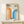 May include: Framed geometric poster with "BAUHAUS 1923" text and curved rainbow stripes in orange and blue gradients, displayed on light wood shelf next to small white wrapped gift with twine and greenery sprig against cream wall