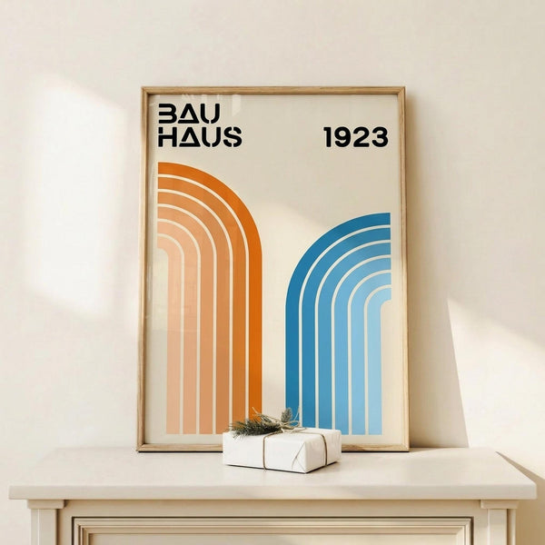 May include: Framed geometric poster with "BAUHAUS 1923" text and curved rainbow stripes in orange and blue gradients, displayed on light wood shelf next to small white wrapped gift with twine and greenery sprig against cream wall