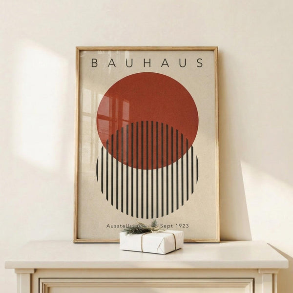 May include: Framed Bauhaus exhibition poster with red circle overlapping black and white striped circle on cream background, displayed on light wood console table next to small wrapped gift with twine and greenery sprig against white wall