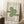 May include: Framed Bauhaus poster with green geometric design featuring flowing curved lines in an abstract pattern, displayed in light wood frame leaning against white wall corner. Scene shows cozy room setting with white coffee cup on wooden block side table and textured beige throw blanket nearby on white fluffy rug.