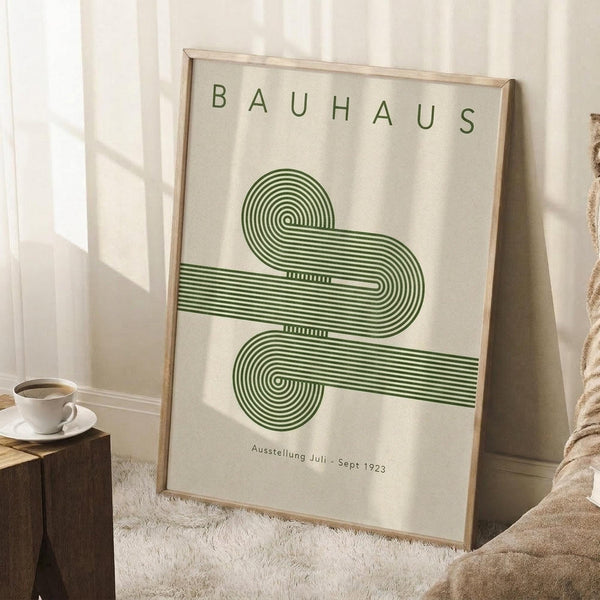 May include: Framed Bauhaus poster with green geometric design featuring flowing curved lines in an abstract pattern, displayed in light wood frame leaning against white wall corner. Scene shows cozy room setting with white coffee cup on wooden block side table and textured beige throw blanket nearby on white fluffy rug.