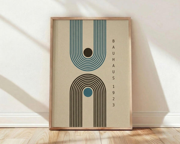May include: Framed geometric poster with cream background featuring two stylized arches in teal and brown with concentric line patterns, black and blue circles as focal points, and vertical text reading "BAUHAUS 1923" on right side. Wooden frame sits on light hardwood floor against white wall with soft natural lighting and shadows.