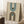 May include: Framed geometric poster with cream background featuring teal and black concentric arches, small dark circle, vertical text reading "BAUHAUS 1923". Wood frame leans against white wall near beige armchair, white coffee cup on wooden side table, textured cream rug below.