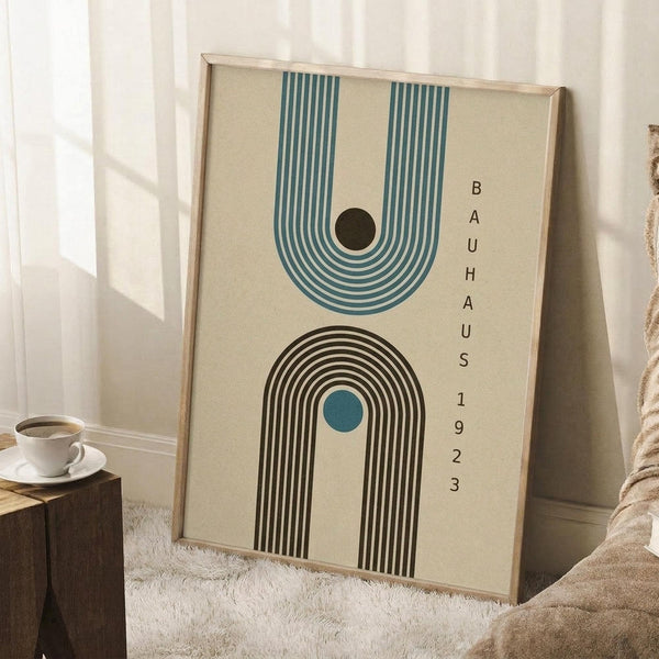 May include: Framed geometric poster with cream background featuring teal and black concentric arches, small dark circle, vertical text reading "BAUHAUS 1923". Wood frame leans against white wall near beige armchair, white coffee cup on wooden side table, textured cream rug below.