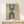 May include: Framed geometric poster with teal and black concentric arches on beige background, displayed on light wood console table with small wrapped gift. The minimalist artwork features two rainbow-shaped designs - one inverted with teal stripes and black dot, one upright with black stripes and teal dot. Text reads "BAUHAUS 1923" vertically. Natural wood frame in clean, modern room setting.