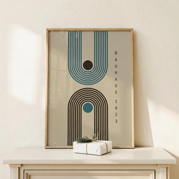 May include: Framed geometric poster with teal and black concentric arches on beige background, displayed on light wood console table with small wrapped gift. The minimalist artwork features two rainbow-shaped designs - one inverted with teal stripes and black dot, one upright with black stripes and teal dot. Text reads "BAUHAUS 1923" vertically. Natural wood frame in clean, modern room setting.