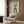 May include: Modern living room with beige armchair displaying framed geometric poster featuring two arch designs - teal and brown striped arches with "BAUHAUS 1923" text, mounted on wood panel wall. Gold floor lamp and dark side table with open book complete the neutral-toned interior scene.