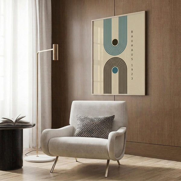 May include: Modern living room with beige armchair displaying framed geometric poster featuring two arch designs - teal and brown striped arches with "BAUHAUS 1923" text, mounted on wood panel wall. Gold floor lamp and dark side table with open book complete the neutral-toned interior scene.