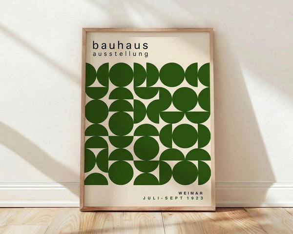 May include: Framed poster with cream background featuring geometric pattern of green circles and semicircles in grid formation. Black text reads "bauhaus ausstellung" at top and "WEIMAR JULI-SEPT 1923" at bottom. Light wood frame sits on wooden floor against white wall with soft window light creating shadows.