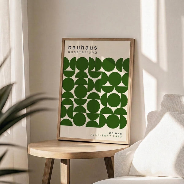 May include: A framed Bauhaus exhibition poster with green geometric circles and semicircles arranged in a grid pattern on cream background, displayed on a light wood round side table next to a beige upholstered chair. Natural sunlight creates soft shadows on white walls, with a plant visible in the corner creating organic shadow patterns.