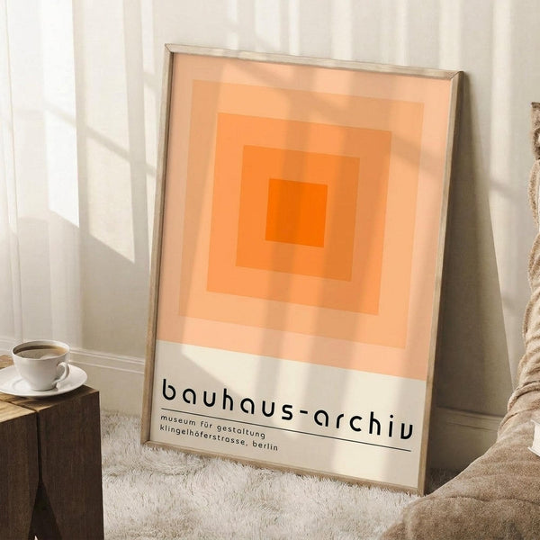 May include: Framed poster with orange gradient squares in Bauhaus style, featuring concentric rectangles from bright orange center fading to pale peach, with "bauhaus-archiv" and German museum text below. Displayed leaning against white wall corner on textured cream rug, next to wooden side table with white coffee cup and saucer, beige throw blanket visible.