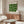 May include: Bauhaus exhibition poster with green geometric circles and text displayed above wooden sideboard in minimalist living room setting. White sculptural chair and glass coffee table visible in foreground. Natural wood tones complement the vintage-inspired wall art in bright, contemporary space with white walls.