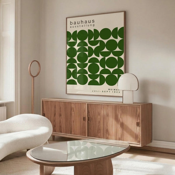May include: Bauhaus exhibition poster with green geometric circles and text displayed above wooden sideboard in minimalist living room setting. White sculptural chair and glass coffee table visible in foreground. Natural wood tones complement the vintage-inspired wall art in bright, contemporary space with white walls.