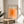 May include: Framed poster with orange gradient squares design and "bauhaus-archiv" text displayed on light wood round side table next to beige upholstered chair. Natural sunlight creates shadows on white wall behind. Green plant leaves visible in corner. Minimalist Scandinavian-style interior setting with warm lighting.