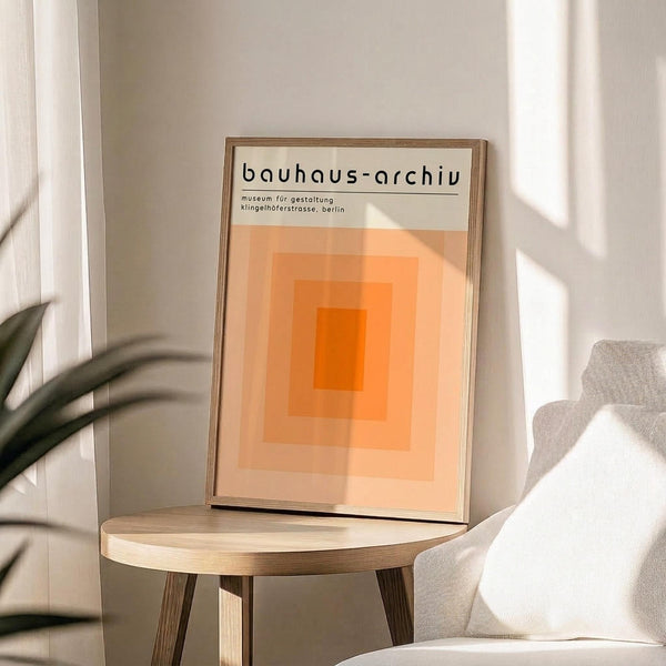 May include: Framed poster with orange gradient squares design and "bauhaus-archiv" text displayed on light wood round side table next to beige upholstered chair. Natural sunlight creates shadows on white wall behind. Green plant leaves visible in corner. Minimalist Scandinavian-style interior setting with warm lighting.