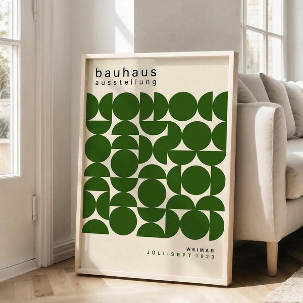 May include: A framed poster with green geometric shapes arranged in a grid pattern on cream background. Black text reads "bauhaus ausstellung" at top and "WEIMAR JULI-SEPT 1923" at bottom. The wooden-framed artwork leans against a white wall corner next to a beige upholstered sofa with light wooden legs. Bright natural light streams through tall white-framed windows onto light hardwood floors.