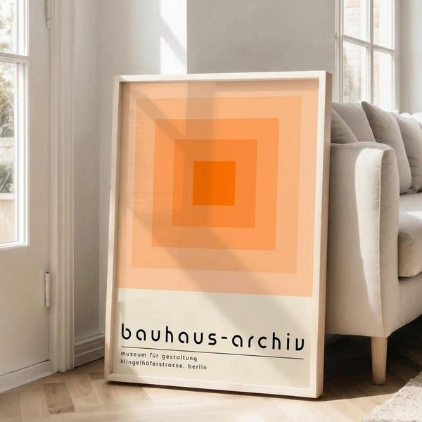 May include: Framed poster with white frame leaning against light gray wall next to beige sofa. Artwork features concentric orange squares in gradient tones from deep orange center to pale peach edges. Black text below reads "bauhaus-archiv" with smaller German text underneath. Bright natural light streams through windows onto hardwood floors with area rug.