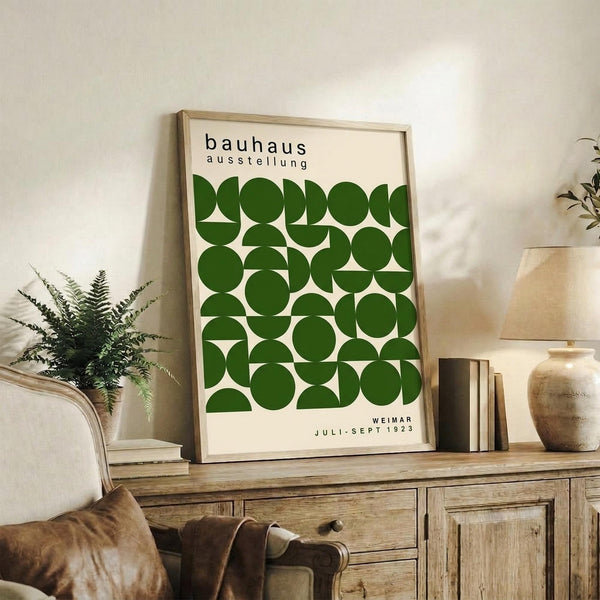 May include: Framed Bauhaus exhibition poster with green geometric circles and semi-circles on cream background, displayed on rustic wooden sideboard alongside fern plant, books, and ceramic vases in warm neutral living room setting