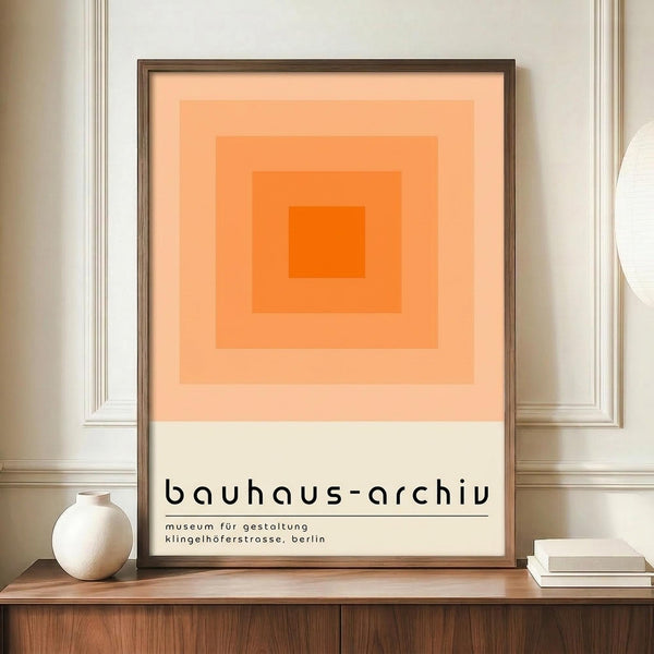 May include: Framed poster with concentric orange squares in gradient shades from deep orange center to pale peach, displayed on wooden sideboard next to white ceramic vase and books. Black text reads "bauhaus-archiv" with German museum details below. Warm beige walls with white molding visible in background.