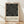 May include: Framed poster with black background featuring white concentric lines forming geometric letter shapes, displayed against white wall on wooden floor. Modern minimalist design with cream-colored lower section containing museum text in simple black typography. Light wooden frame with natural shadows cast across the scene.