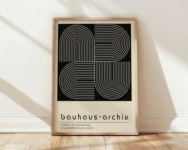 May include: Framed poster with black background featuring white concentric lines forming geometric letter shapes, displayed against white wall on wooden floor. Modern minimalist design with cream-colored lower section containing museum text in simple black typography. Light wooden frame with natural shadows cast across the scene.