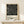 May include: Framed Bauhaus-style poster with geometric black and white line design forming abstract letters, displayed on light wood console table beside small wrapped gift with twine and evergreen sprig, against cream wall in minimalist room setting