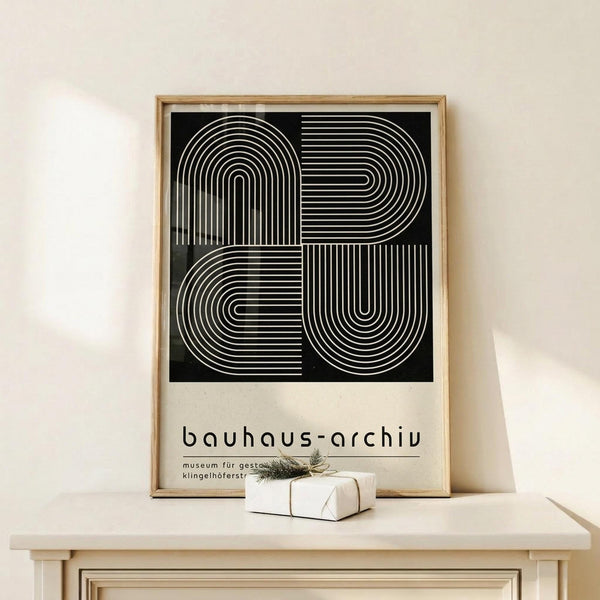 May include: Framed Bauhaus-style poster with geometric black and white line design forming abstract letters, displayed on light wood console table beside small wrapped gift with twine and evergreen sprig, against cream wall in minimalist room setting