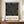 May include: Framed Bauhaus-style poster with black background featuring white geometric line art forming curved and angular shapes in four quadrants. Text reads "bauhaus-archiv museum für gestaltung klingelhöferstrasse, berlin" below the design. Displayed on wooden sideboard next to cream ceramic vase and stacked books in room with beige walls and white molding.