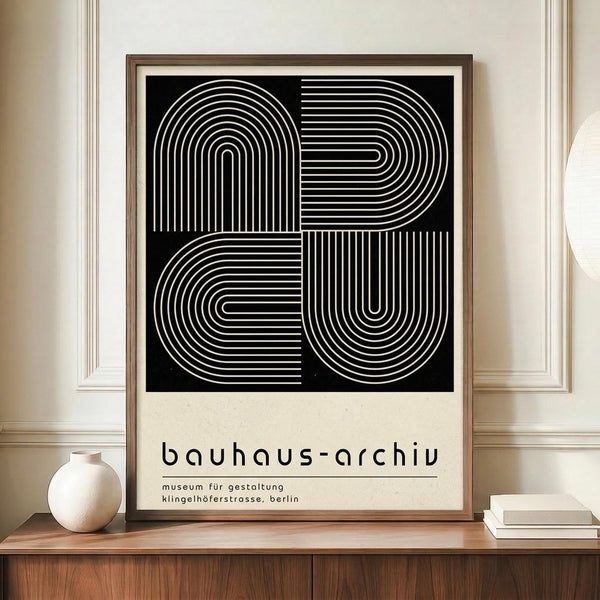 May include: Framed Bauhaus-style poster with black background featuring white geometric line art forming curved and angular shapes in four quadrants. Text reads "bauhaus-archiv museum für gestaltung klingelhöferstrasse, berlin" below the design. Displayed on wooden sideboard next to cream ceramic vase and stacked books in room with beige walls and white molding.