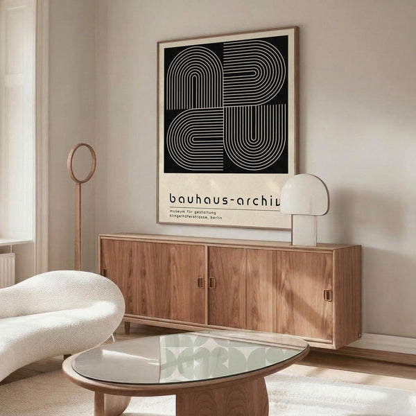 May include: Black and white geometric poster with curved linear patterns forming abstract shapes, displayed in wooden frame above mid-century modern walnut credenza. Room features cream upholstered chair, glass-top oval coffee table, modern white table lamp, and neutral beige walls with natural lighting.