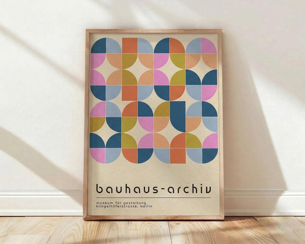 May include: Framed Bauhaus-style poster with geometric pattern of colorful circles and squares in blues, oranges, pinks, and yellows on cream background, displayed on light wooden floor against white wall with soft natural lighting and shadows