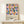 May include: Framed geometric poster with colorful circles and squares in pink, blue, orange, and olive green pattern displayed on light wooden console table against white wall. Small wrapped white gift box with twine and pine sprig sits nearby. Modern Scandinavian-style room setting with natural wood frame.