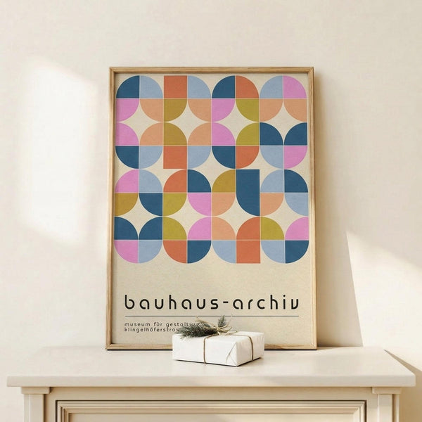 May include: Framed geometric poster with colorful circles and squares in pink, blue, orange, and olive green pattern displayed on light wooden console table against white wall. Small wrapped white gift box with twine and pine sprig sits nearby. Modern Scandinavian-style room setting with natural wood frame.