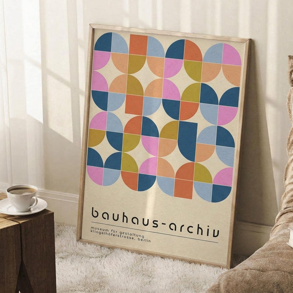 May include: Framed geometric art print with colorful overlapping circles in pink, blue, orange, and yellow tones on cream background, leaning against white wall next to wooden side table with coffee cup, placed on textured white rug with cozy knit blanket nearby in soft natural lighting.