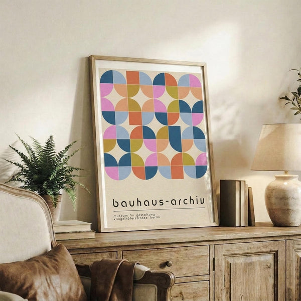 May include: Framed geometric art print with colorful circles and semicircles in pink, blue, orange, and green displayed on rustic wooden sideboard. Room setting features neutral armchair, potted fern, ceramic vases, and stacked books creating modern minimalist home decor scene with warm natural lighting.
