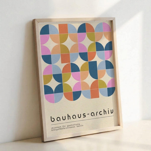 May include: Framed poster with geometric pattern of colorful overlapping circles in blue, pink, orange, yellow and coral tones arranged in a grid. Text reads "bauhaus-archiv" with smaller German text below. Light wooden frame against white wall, natural lighting casting soft shadows.