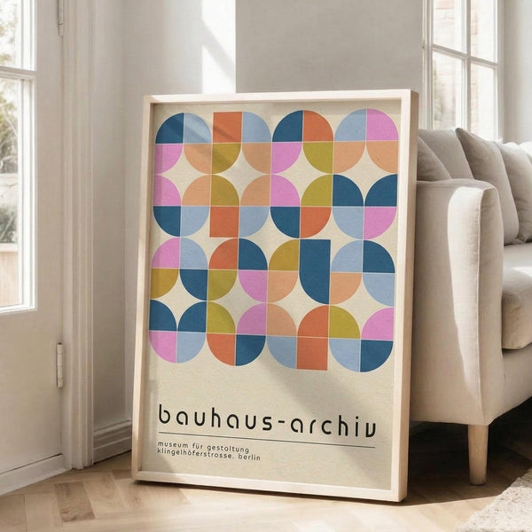 May include: Framed geometric art print leaning against white wall beside beige sofa in bright living room. Colorful poster features overlapping circles and squares in pink, blue, orange, and yellow arranged in grid pattern. Black text below reads "bauhaus-archiv" with museum details. Natural wood frame, hardwood floors, large windows with natural lighting.