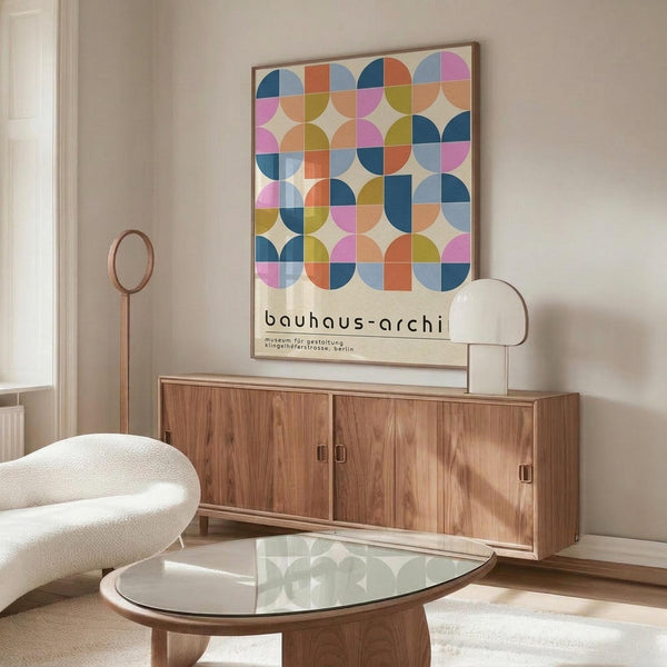 May include: Modern living room with walnut credenza displaying colorful geometric Bauhaus poster featuring overlapping circles in pink, blue, orange, and green. Cream upholstered chair, brass floor lamp, and oval glass coffee table complete the mid-century inspired space with white walls and natural lighting.