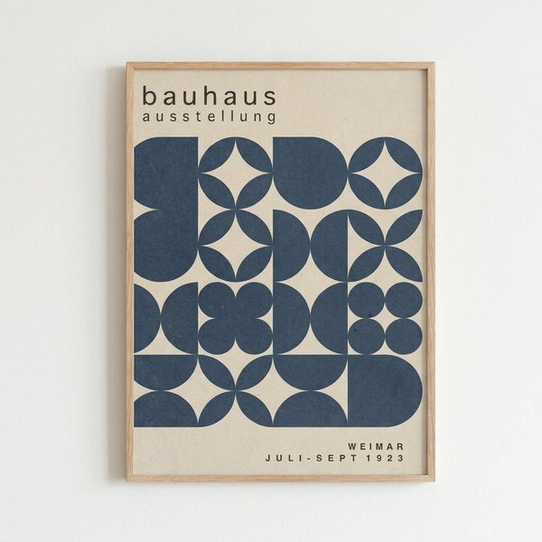 May include: Framed Bauhaus exhibition poster mounted on white wall. Design features geometric pattern of navy blue circles and semicircles creating diamond and quatrefoil shapes on cream background. Text reads "bauhaus ausstellung" at top and "WEIMAR JULI-SEPT 1923" at bottom. Simple light wood frame. Clean minimalist wall display showcasing vintage German design school aesthetic with bold geometric forms.