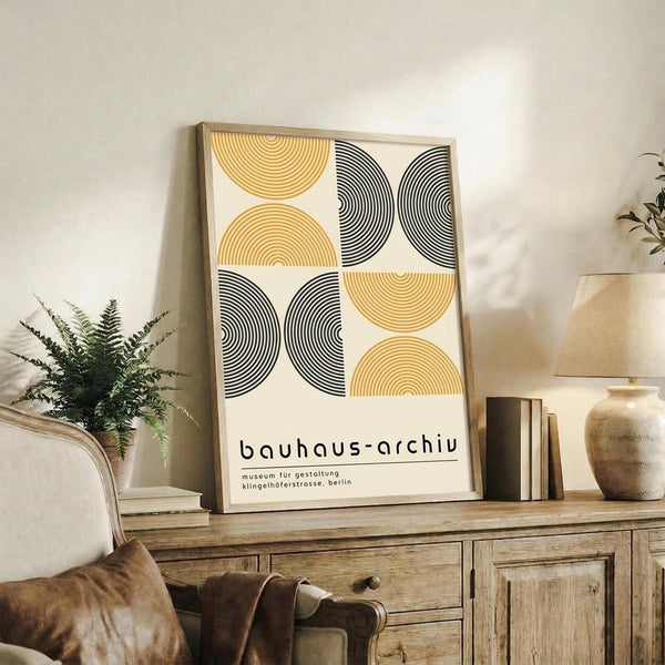 May include: Framed Bauhaus-style poster with geometric semicircles in yellow and black concentric lines on cream background, displayed on rustic wooden sideboard alongside fern plant, books, ceramic vase, and brown leather chair in warm neutral living room setting