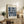 May include: Framed geometric poster with blue circles and overlapping patterns creating star-like shapes on cream background, displayed on rustic wooden sideboard alongside potted fern, ceramic vases, and stacked books in neutral-toned living room setting with beige upholstered furniture
