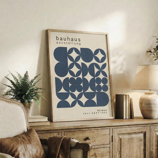 May include: Framed geometric poster with blue circles and overlapping patterns creating star-like shapes on cream background, displayed on rustic wooden sideboard alongside potted fern, ceramic vases, and stacked books in neutral-toned living room setting with beige upholstered furniture
