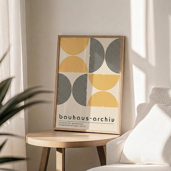 May include: Framed geometric poster with yellow and black semicircular patterns arranged in grid format, displayed on light wood round side table next to beige upholstered chair. Natural lighting creates soft shadows on neutral wall background with plant leaves visible in corner.