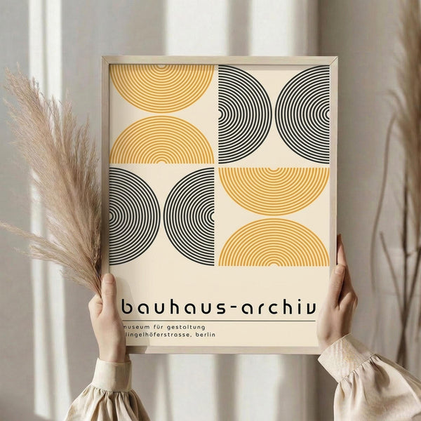 May include: Hands holding a framed poster with geometric semicircular patterns in golden yellow and black concentric lines against cream background, displayed next to dried pampas grass in neutral interior setting with natural lighting and beige curtains