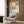 May include: Geometric poster with yellow and black semicircular patterns hung on wood-paneled wall above beige upholstered armchair with patterned throw pillow. Modern brass floor lamp and dark side table with open book visible in neutral-toned room setting with curtains and wood flooring.