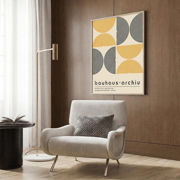 May include: Geometric poster with yellow and black semicircular patterns hung on wood-paneled wall above beige upholstered armchair with patterned throw pillow. Modern brass floor lamp and dark side table with open book visible in neutral-toned room setting with curtains and wood flooring.