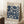 May include: A framed Bauhaus exhibition poster with blue geometric circular patterns on cream background, leaning against white wall. Wood frame, displayed with coffee cup on wooden block and beige knit throw on white textured rug. Natural lighting creates soft shadows.