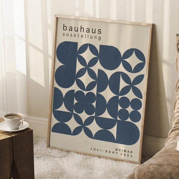 May include: A framed Bauhaus exhibition poster with blue geometric circular patterns on cream background, leaning against white wall. Wood frame, displayed with coffee cup on wooden block and beige knit throw on white textured rug. Natural lighting creates soft shadows.